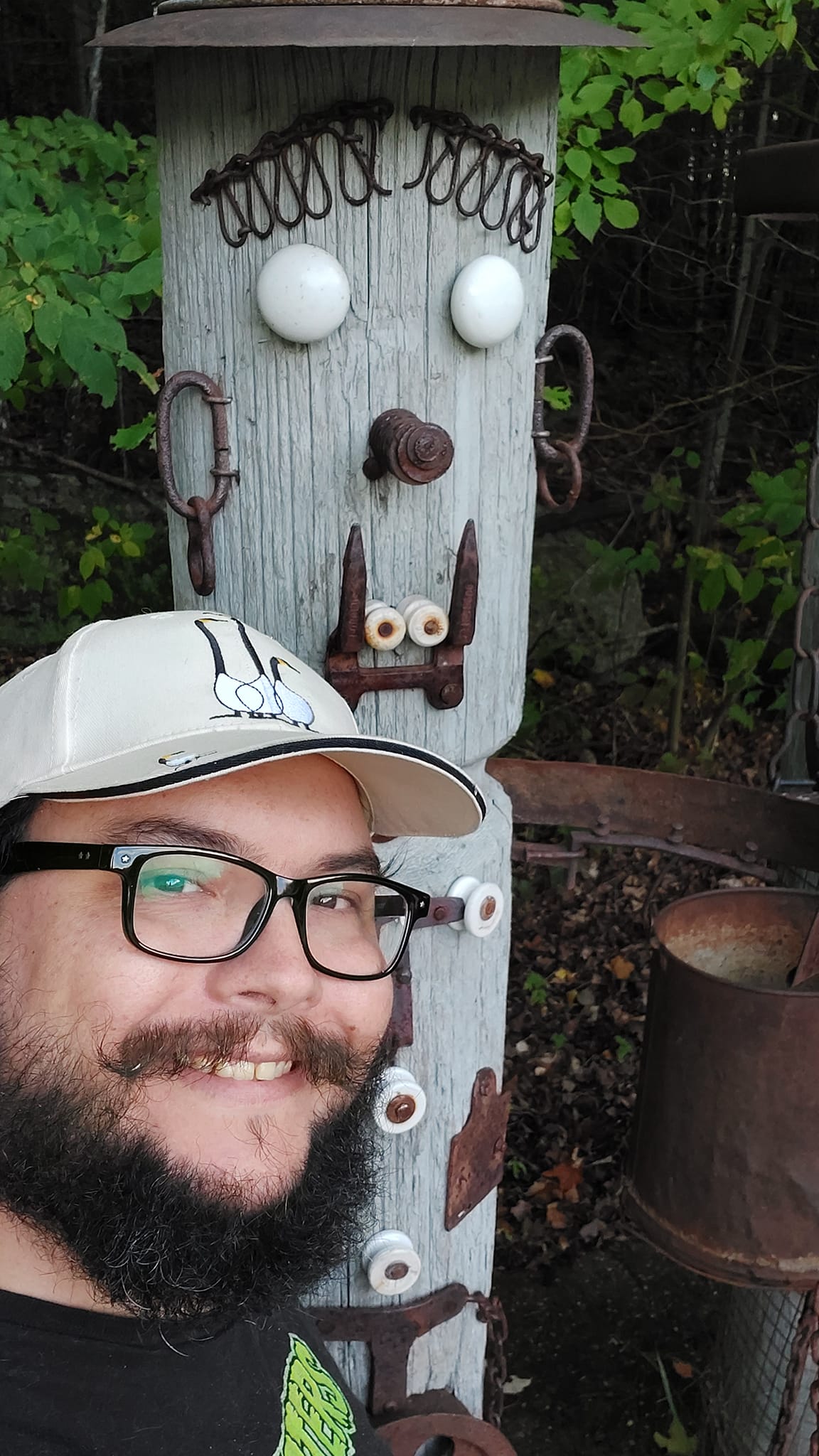 A man with short hair and bushy facial hair stands in front of a sculpture primarily made of wood with metal and other objects afixed to it, it has a face and is supposed to be reminiscent of a settler, it is taller than the man